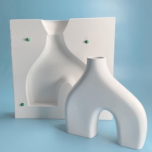 May include: A white ceramic vase with an abstract, asymmetrical design. The vase is shown next to its mold, which is also white.