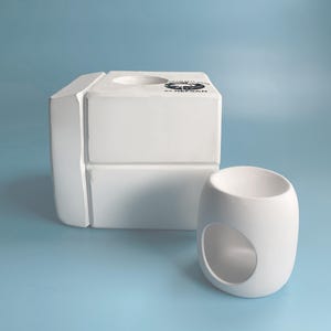 May include: White ceramic wax melt warmer with a square base and a round top. The warmer has a small hole in the top for a tea light candle. The warmer is sitting on a light blue surface.