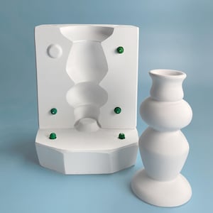 May include: A white ceramic mould for making a vase with a round base and a bulbous body. The mould has five green circles on the sides. Next to the mould is a white ceramic vase with a round base and a bulbous body.
