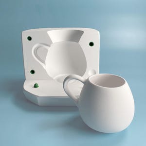 Puede incluir: Una taza de cerámica blanca con cuerpo redondeado y asa. La taza está junto a un molde blanco para hacer la taza.