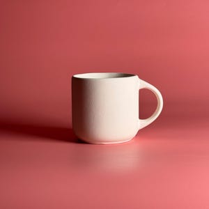 May include: A cream-colored ceramic mug with a rounded base and a curved handle. The mug is set against a solid red-pink background. The mug's simple design and neutral color make it suitable for various beverages.