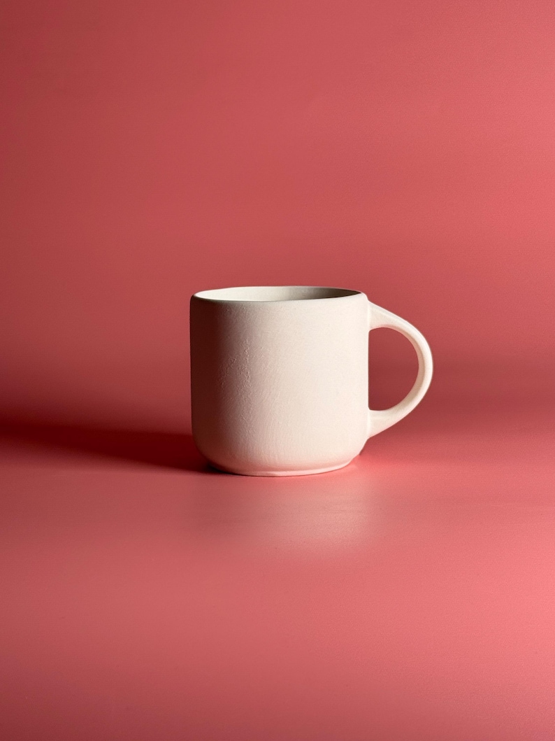 May include: A cream-colored ceramic mug with a rounded base and a curved handle. The mug is set against a solid red-pink background. The mug's simple design and neutral color make it suitable for various beverages.