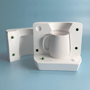 Pode incluir: Um molde de caneca de cer&acirc;mica branca com um orif&iacute;cio de ventila&ccedil;&atilde;o verde. O molde est&aacute; aberto e a forma da caneca &eacute; vis&iacute;vel.