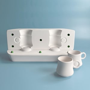 Op de afbeelding: Witte keramische mal voor het maken van drie bekers, met twee afgewerkte witte keramische bekers met handvatten.