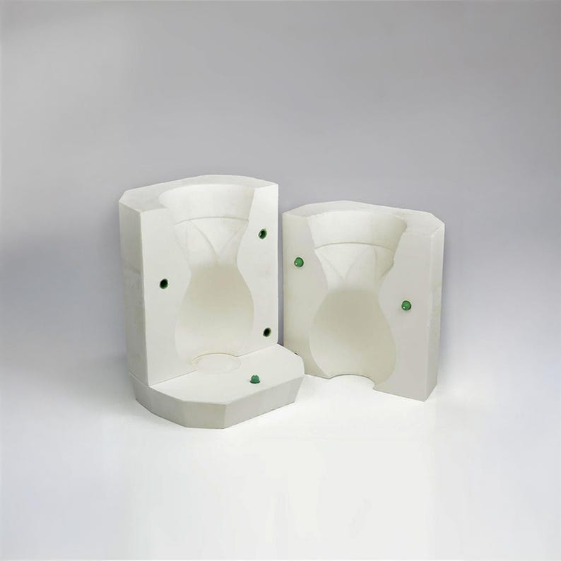 May include: Two white ceramic molds for making a vase or pitcher. The molds are open and show the inside of the vase shape. The molds have small green circles on the sides.