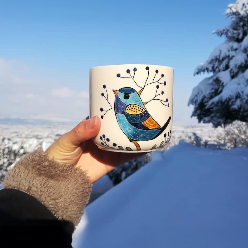 May include: A white ceramic mug featuring a hand-painted blue bird design with orange and yellow accents. The bird is perched on a branch with black berries. The mug is held against a snowy winter landscape.