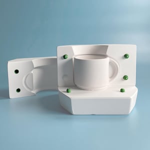 May include: A white ceramic mug sits inside a white mold, with a second mold piece to the left. The molds have green accents. The mug has a curved handle and a smooth, matte finish. The background is a solid light blue.