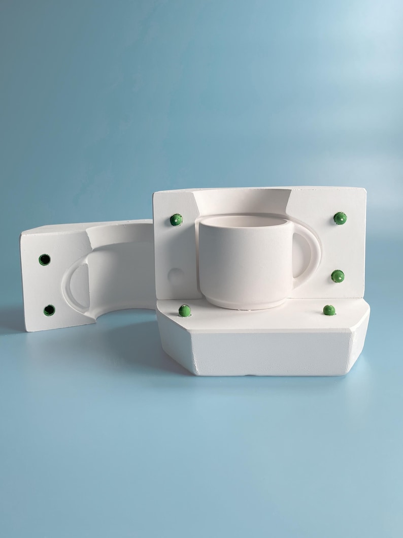 May include: A white ceramic mug sits inside a white mold, with a second mold piece to the left. The molds have green accents. The mug has a curved handle and a smooth, matte finish. The background is a solid light blue.