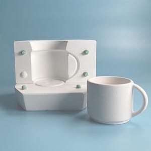 以下が含まれることがあります： 白い取っ手付きのセラミックマグが、緑色のアクセントが付いた白い型枠の隣に置かれています。マグはシンプルで円筒形のデザインです。型枠は同じ形のマグを作成するように設計されています。背景は単色の水色です。