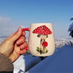 Pode incluir: Uma caneca de cer&acirc;mica branca com um desenho de cogumelo vermelho e branco. A caneca tem uma al&ccedil;a vermelha e &eacute; decorada com manchas brancas.