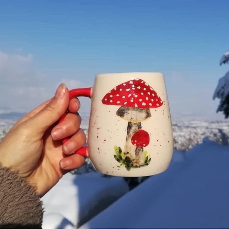 Pode incluir: Uma caneca de cer&acirc;mica branca com um desenho de cogumelo vermelho e branco. A caneca tem uma al&ccedil;a vermelha e &eacute; decorada com manchas brancas.