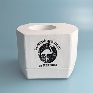 May include: A white, octagonal ceramic holder with a circular opening at the top. The front features a black circular logo with a flamingo and the text "ceramingo.com BY REFSAN". The holder is set against a light blue background.