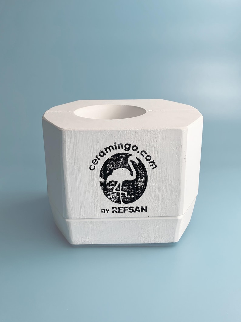 May include: A white, octagonal ceramic holder with a circular opening at the top. The front features a black circular logo with a flamingo and the text "ceramingo.com BY REFSAN". The holder is set against a light blue background.
