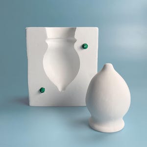 May include: A white ceramic mould for making a vase, with a green gemstone accent. A finished vase made from the mould is shown next to it.