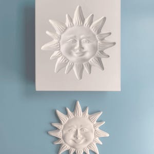 以下が含まれることがあります： 笑顔の顔と光線を備えた白いセラミック製の太陽の型。型は薄い青色の背景の上にあります。