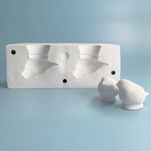 Op de afbeelding: Een witte keramische mal met twee vogelvormige holtes en twee kleine, witte keramische vogelbeeldjes. De mal heeft groene stippen en een glad oppervlak. De achtergrond is lichtblauw.