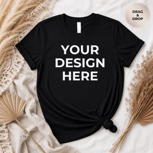 Puede incluir: Camiseta negra con el texto "YOUR DESIGN HERE" en letras blancas. La camiseta tiene cuello redondo y mangas cortas. La camiseta está anudada en la parte inferior. El fondo incluye plantas secas y una tela de color neutro.