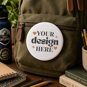 Peut inclure: Un badge rond blanc avec le texte gris foncé "YOUR design HERE" écrit dans une police serif grasse, agrémenté de deux petits cœurs dorés et de deux étoiles dorées à quatre branches, fixé sur la poche avant d'un sac à dos en toile vert olive.