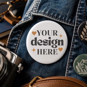 May include: A circular white pin-back button featuring the dark gray text "YOUR design HERE" in a bold serif font. The design is accented with two small gold-colored hearts and two four-pointed star icons, creating a clean and simple graphic display.