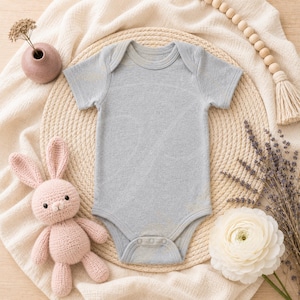 Peut inclure: Un body de bébé gris clair à manches courtes et fermetures à boutons-pression, présenté sur un tapis tissé. Un jouet lapin rose crocheté, une fleur blanche et des éléments décoratifs entourent le body. La scène est posée sur un tissu crème.