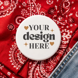 May include: A white button with the text "YOUR design HERE" in a stylish font, accented with gold hearts and stars. The button is set against a red paisley bandana and denim fabric.