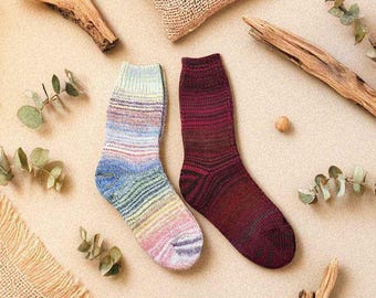 May include: Two pairs of colorful knitted socks. One pair features a gradient of blue, green, yellow, and pink. The other pair is a deep red with darker red stripes. The socks are displayed on a beige surface with decorative elements.