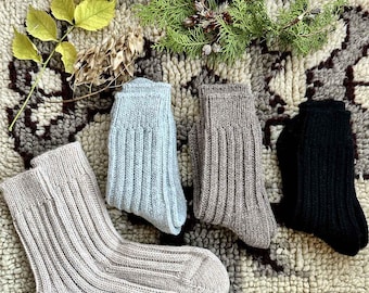 May include: Four pairs of ribbed knit socks in various colors: light gray, light blue, taupe, and black. The socks are arranged on a patterned rug with sprigs of greenery and dried foliage.