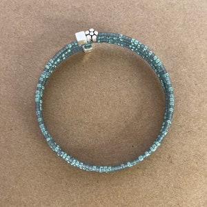 Puede incluir: Un brazalete de cuentas de plata y turquesa con un cierre de plata. El brazalete está hecho de pequeñas cuentas redondas y tiene un diámetro de unos 18 cm.