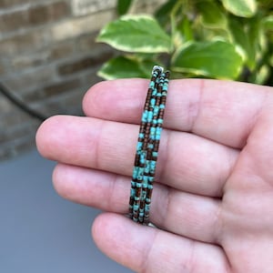 Könnte beinhalten: Nahaufnahme eines mehrreihigen Armbands aus kleinen türkisfarbenen und braunen Perlen. Das Armband ist um eine Hand gewickelt.