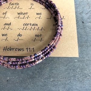 Peut inclure: Gros plan sur un bracelet en perles brunes et violettes avec une carte qui dit "la foi n'est pas de ce que nous faisons certain Hébreux 11:1".