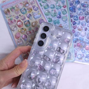 May include: A clear phone case with a grid of decorative charms, including bows and hearts, is held in front of several sticker sheets. The sticker sheets feature various cute, colorful designs, such as animals and flowers. The stickers are in shades of blue, pink, and purple.
