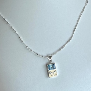 May include: A silver-toned necklace featuring a pendant shaped like a handheld gaming console. The pendant includes a rectangular blue gemstone screen and two small circular clear stones representing buttons, suspended from a delicate beaded chain.