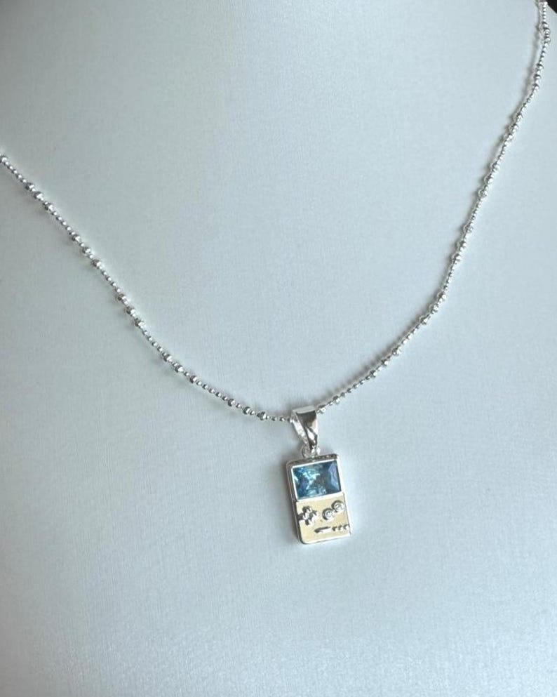 May include: A silver-toned necklace featuring a pendant shaped like a handheld gaming console. The pendant includes a rectangular blue gemstone screen and two small circular clear stones representing buttons, suspended from a delicate beaded chain.