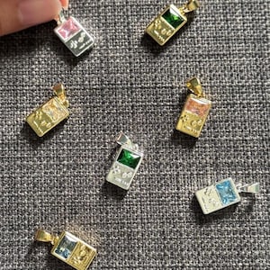 May include: A collection of seven rectangular pendants shaped like handheld video game consoles. Each pendant features a central rectangular gemstone in colors including pink, green, clear, orange, and blue, set in either gold or silver-toned metal.