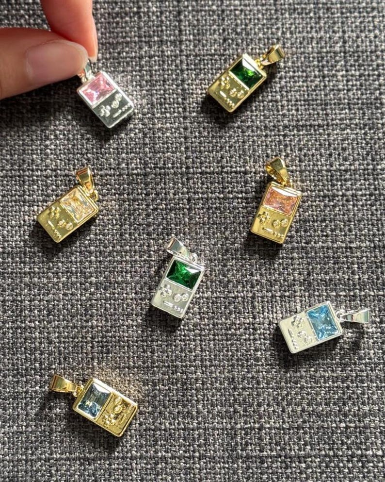 May include: A collection of seven rectangular pendants shaped like handheld video game consoles. Each pendant features a central rectangular gemstone in colors including pink, green, clear, orange, and blue, set in either gold or silver-toned metal.