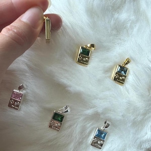 May include: A collection of small handheld game console pendants in gold and silver tones. Each pendant features a rectangular gemstone in pink, green, or blue, with engraved button details on the lower portion and a small metal bail at the top.