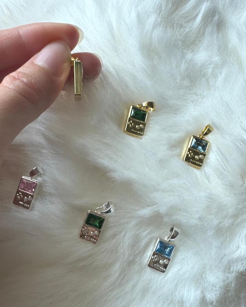 May include: A collection of small handheld game console pendants in gold and silver tones. Each pendant features a rectangular gemstone in pink, green, or blue, with engraved button details on the lower portion and a small metal bail at the top.