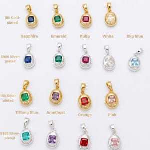 May include: A collection of Gemagotchi pendants arranged in rows, featuring 18k gold-plated and S925 silver-plated settings. Each oval pendant contains a square-cut gemstone in various colors including sapphire, emerald, ruby, white, sky blue, tiffany blue, amethyst, orange, and pink, accented with three small clear stones at the base.
