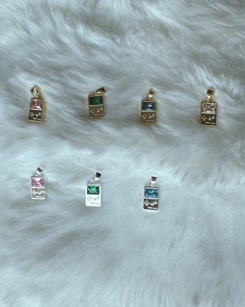 May include: A collection of seven rectangular pendants shaped like handheld video game consoles. Four pendants are gold-toned with pink, green, dark blue, and clear stones. Three pendants are silver-toned with pink, green, and blue stones.
