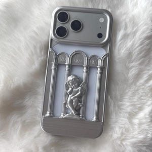 May include: Silver phone case with a classical architectural design. The case features a relief of an angel and columns. The phone case is silver and has a white background.