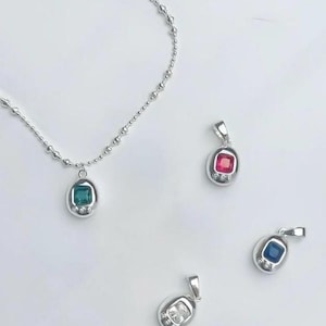 Puede incluir: Un collar de plata con un colgante de piedra verde y tres colgantes de piedras preciosas adicionales en rojo, azul y transparente. Cada colgante tiene una forma ovalada pequeña con un engaste de bisel y un pequeño lazo para una cadena.