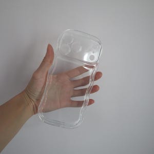 May include: Clear, transparent phone case with a wavy, irregular edge. The case has cutouts for the phone's camera lenses and buttons. The case is held in a hand against a plain white background.