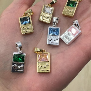 May include: A collection of seven metal pendants shaped like handheld video game consoles. Each pendant features a rectangular gemstone in colors including green, blue, clear, and pink, set into gold-tone or silver-tone metal frames with button details.