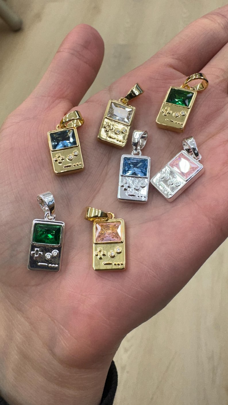 May include: A collection of seven metal pendants shaped like handheld video game consoles. Each pendant features a rectangular gemstone in colors including green, blue, clear, and pink, set into gold-tone or silver-tone metal frames with button details.