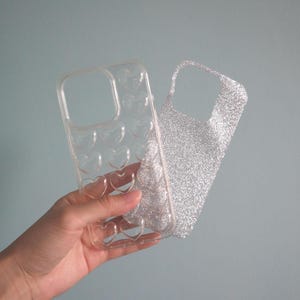 May include: Clear phone case with heart-shaped cutouts and a glittery silver insert. The case is held up against a light blue background. The case is designed to protect the phone.