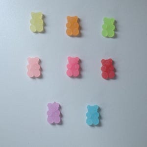 May include: A collection of nine gummy bear candies in various colors, including yellow, orange, green, pink, red, purple, and blue. The gummy bears are arranged on a white surface, each with a translucent appearance.