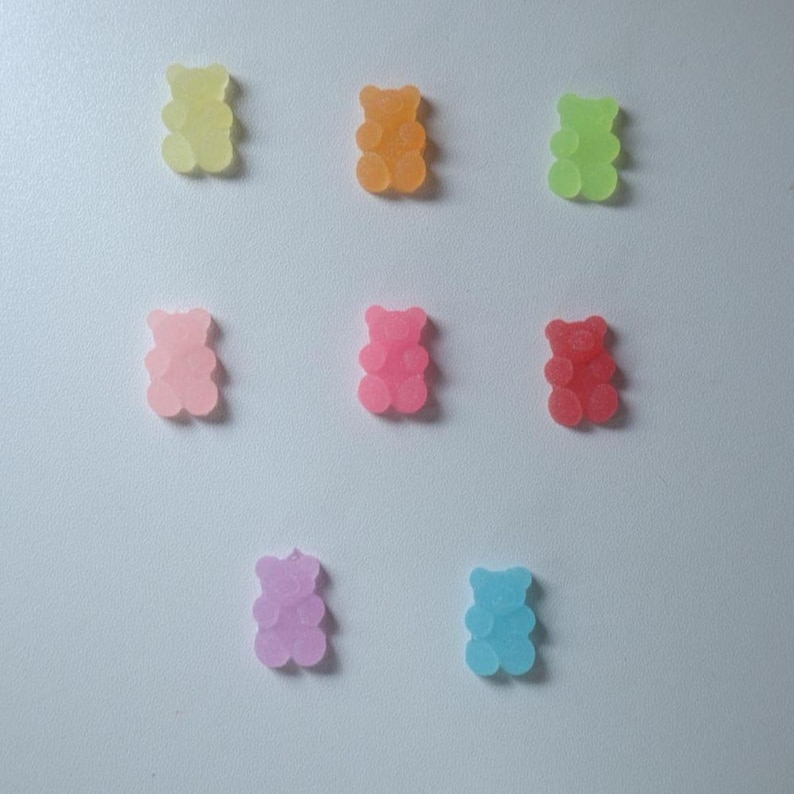 May include: A collection of nine gummy bear candies in various colors, including yellow, orange, green, pink, red, purple, and blue. The gummy bears are arranged on a white surface, each with a translucent appearance.