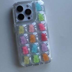 May include: Clear phone case with a colorful gummy bear design. The case features a grid pattern filled with various colored gummy bear shapes, including blue, green, pink, orange, and purple. The phone's camera lenses are visible at the top.