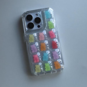 May include: Clear phone case with colorful gummy bear accents. The case features a grid pattern with individual compartments, each holding a gummy bear in various colors, including pink, green, orange, and blue. The case is designed to fit a smartphone.