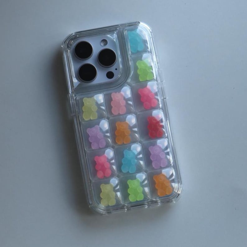 May include: Clear phone case with colorful gummy bear accents. The case features a grid pattern with individual compartments, each holding a gummy bear in various colors, including pink, green, orange, and blue. The case is designed to fit a smartphone.